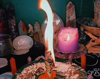 May include: A lit candle with a tall flame sits on a decorative plate surrounded by crystals. A pink candle and other crystals are in the background. The plate has a green rim and is covered in herbs.