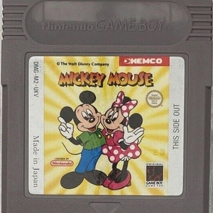 May include: A gray Nintendo Game Boy cartridge featuring the game "Mickey Mouse." The label shows Mickey and Minnie Mouse, with the text "The Walt Disney Company" and "Made in Japan." The cartridge is a classic video game accessory.