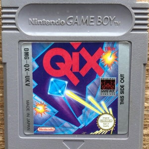 Peut inclure: Une cartouche grise Nintendo Game Boy pour le jeu QIX. La cartouche a une étiquette colorée avec le titre du jeu en rouge, des formes géométriques et le logo Nintendo. L'étiquette comprend également le texte "MADE IN JAPAN".