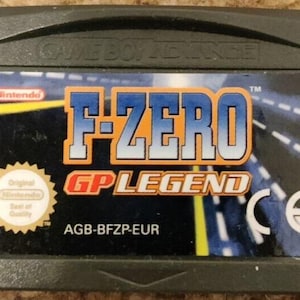Può includere: Cartuccia per Game Boy Advance per F-Zero GP Legend. L'etichetta presenta il titolo del gioco in blu e arancione, con uno sfondo di scena di corsa. La cartuccia è grigio scuro, con il logo Nintendo e un sigillo di qualità.