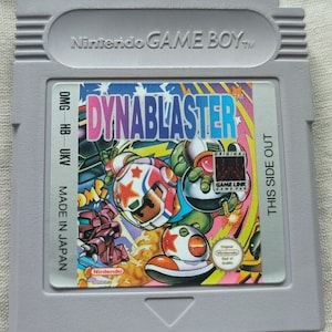 Könnte beinhalten: Eine graue Nintendo Game Boy-Cartridge für das Spiel Dynablaster. Die Cartridge zeigt farbenfrohe Grafiken mit dem Titel des Spiels in Pink und Lila. Die Cartridge ist mit "Made in Japan" und "This Side Out" gekennzeichnet.