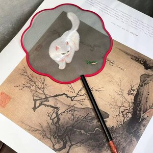 May include: A decorative hand fan with a white cat illustration on a mesh surface, framed by a red scalloped edge. The fan has a long black handle with a wood-grain finish at the base. The background features an Asian-style painting.