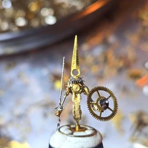 May include: A miniature figurine constructed from metallic watch parts, featuring a tall pointed head, a slender arm holding a thin rod, and a circular gear acting as a shield. The figure is mounted on a small white base with a black bottom.