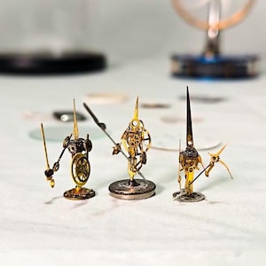 May include: Three miniature steampunk warrior figurines made from watch components. The figures are gold and silver, with detailed gears and cogs. Each holds a weapon, like a sword or spear, and stands on a small base.