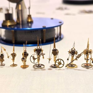 May include: A collection of miniature brass and gold-toned mechanical sculptures. Each sculpture features intricate gears, levers, and other mechanical components. A blue and silver-toned circular base is in the background. The sculptures are arranged in a row.