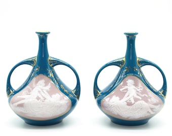 May include: Pair of teal blue ceramic vases with gold accents and white relief sculptures of a woman and a cherub. The vases have two handles on each side.