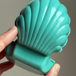 May include: A turquoise plastic container shaped like a scallop shell with a hinged lid. The base of the object features the text MADE IN U.S.A. molded into the surface. The shell design has vertical ridges and a smooth, glossy finish throughout.