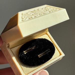May include: An open ivory-coloured jewellery box with a floral pattern on the lid. Inside, a black velvet insert with two slots sits on a gold-coloured fabric lining. The box is held in a person's hand.
