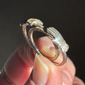 May include: A silver ring featuring two hands clasped together. The ring has an open design with intricate detailing on the band and the hands. The ring is shown in a close-up shot, highlighting its delicate craftsmanship.