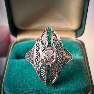 May include: An antique-style ring with a central square-cut diamond and emerald accents. The ring features intricate filigree detailing in a silver or white gold setting, presented in a green velvet ring box.
