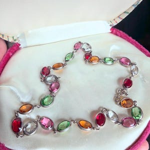 Op de afbeelding: Een zilverkleurige armband met ovale edelstenen in verschillende kleuren, waaronder rood, oranje, groen, roze en helder. De armband wordt gepresenteerd in een roze fluwelen sieradendoos met een crèmekleurig satijnen interieur.
