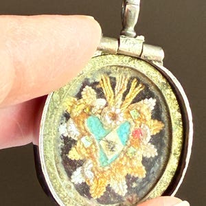 Pode incluir: Pingente redondo e antigo com um design detalhado. Apresenta um coração turquesa central, rodeado por elementos florais dourados e brancos. O pingente tem uma moldura de metal prateado e uma pequena argola na parte superior.