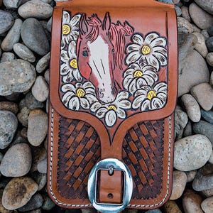 May include: A brown leather saddle shop accessory with a horse head and daisy design. The accessory has a woven pattern and a silver buckle. The text "Atomic Buckaroo Saddle Shop" is visible.