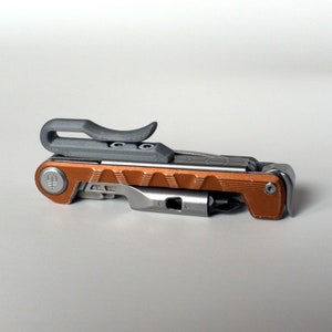 Könnte beinhalten: Ein Multitool mit kupferfarbenem Griff und einem grauen Clip. Das Werkzeug verfügt über eine Vielzahl von Klingen und Werkzeugen, darunter einen Schraubendreher, ein Messer und eine Zange.