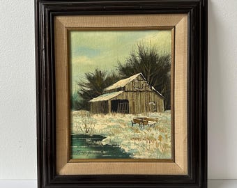 May include: An oil painting of a rustic barn in a winter landscape. The artwork features a brown barn with a snow-covered roof, set against a backdrop of bare trees and a pale blue sky. The painting is framed in a dark brown wooden frame.