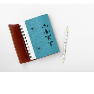 May include: A teal blue leather bound notebook with a spiral binding. The front cover features a series of black stick figures performing different weightlifting exercises. The notebook is open to a blank page and a white pen with black polka dots is lying next to it.
