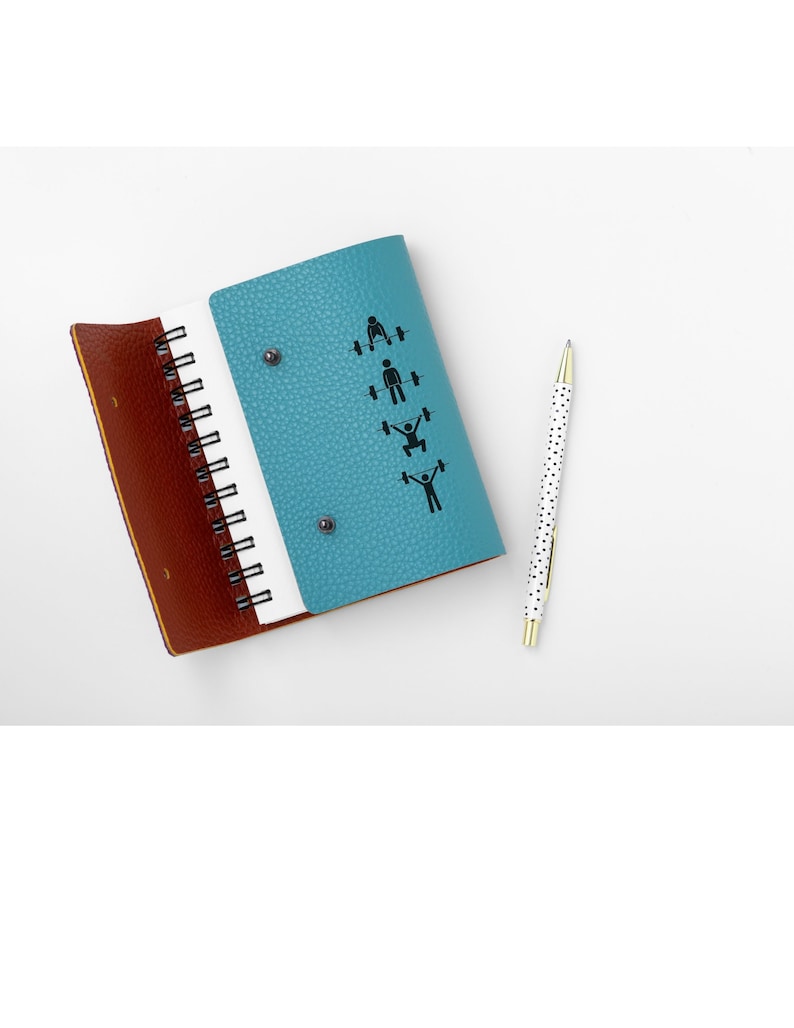 May include: A teal blue leather bound notebook with a spiral binding. The front cover features a series of black stick figures performing different weightlifting exercises. The notebook is open to a blank page and a white pen with black polka dots is lying next to it.