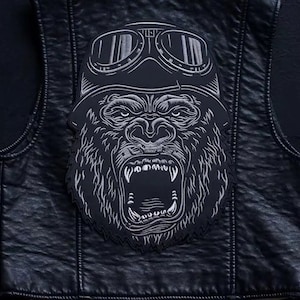 Puede incluir: Un chaleco de cuero negro para motocicleta con un parche bordado en plata de un gorila con gafas. La boca del gorila está abierta y se le ven los dientes.