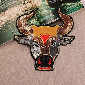 May include: A gold, brown, black, and red sequined bull head applique. The bull's eyes are black with white highlights. The bull's nose is black with red nostrils. The bull's horns are gold.