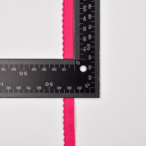 以下が含まれることがあります： 定規で測定された明るいピンクの波状のトリムのクローズアップ。トリムは約1.2cmの幅です。