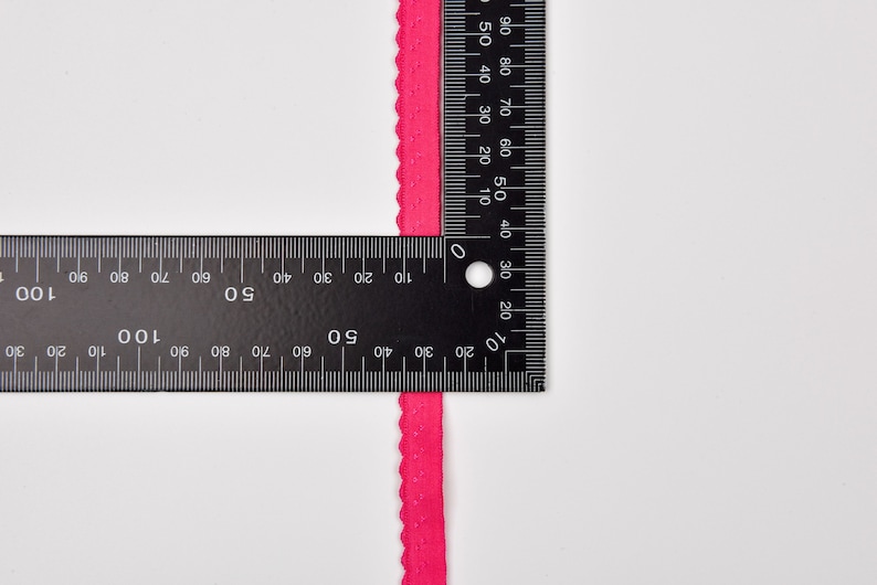 以下が含まれることがあります： 定規で測定された明るいピンクの波状のトリムのクローズアップ。トリムは約1.2cmの幅です。