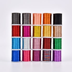 Puede incluir: Una colección de 20 carretes de hilo metálico en varios colores, incluyendo rojo, azul, verde, dorado, plateado y morado. Los carretes están dispuestos en cuatro filas de cinco, y cada carrete tiene una base de plástico blanco.