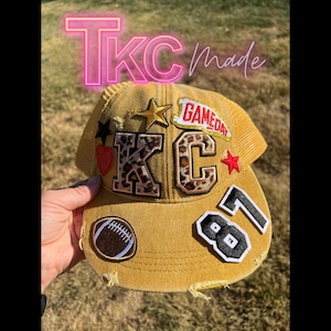 May include: A mustard yellow distressed trucker hat featuring various patches including leopard print letters K and C, a red heart, a gold star, a football, the number 87 in black and white, and a banner with the red text GAMEDAY.