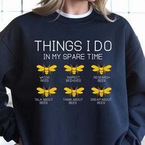 May include: Navy blue sweatshirt with white text that reads "THINGS I DO IN MY SPARE TIME." Below the text are six yellow bee graphics with corresponding text: Watch Bees, Inspect Beehives, Research Bees, Talk About Bees, Think About Bees, and Dream About Bees.