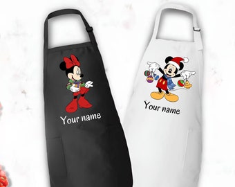 May include: Two aprons, one black and one white, each featuring a Disney character. The black apron has Minnie Mouse in a holiday outfit, and the white apron has Mickey Mouse in a Santa hat. Both aprons have the text "Your name".