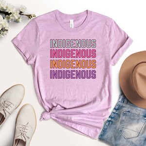 May include: A pink t-shirt with the word "INDIGENOUS" repeated four times in a stacked design. The text is in a rainbow color scheme with a white outline.