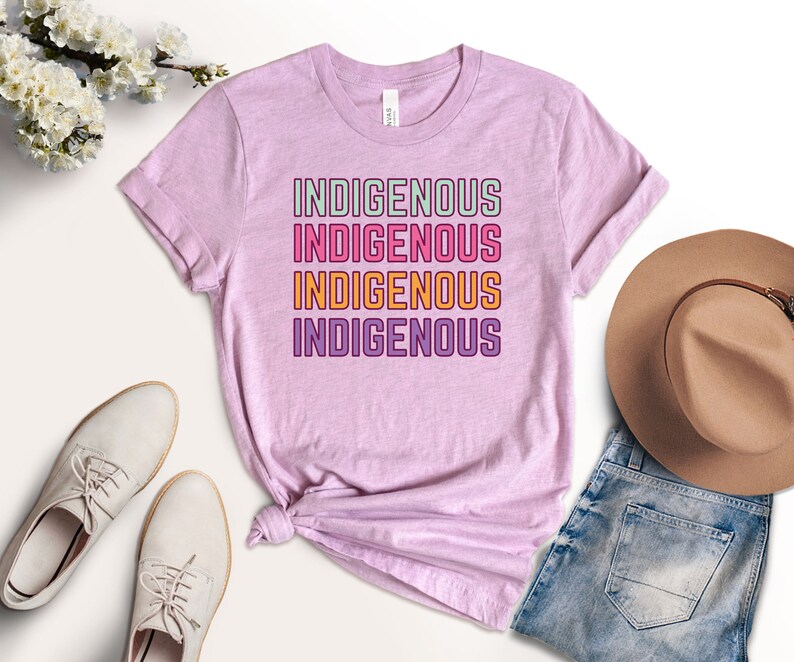 May include: A pink t-shirt with the word "INDIGENOUS" repeated four times in a stacked design. The text is in a rainbow color scheme with a white outline.