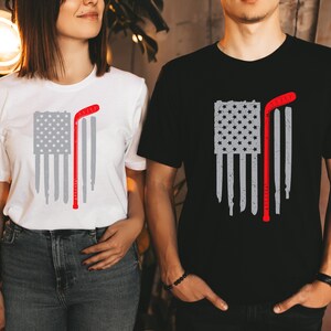 Puede incluir: Dos camisetas con un dise&ntilde;o de bandera estadounidense desgastada y un palo de hockey. La bandera es gris y el palo de hockey es rojo. Una camiseta es blanca y la otra es negra.