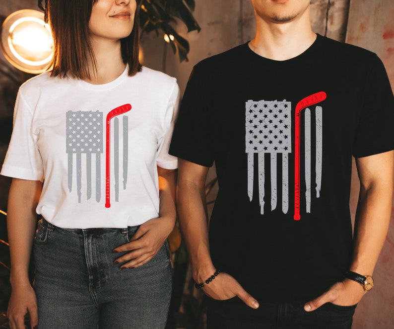 Puede incluir: Dos camisetas con un dise&ntilde;o de bandera estadounidense desgastada y un palo de hockey. La bandera es gris y el palo de hockey es rojo. Una camiseta es blanca y la otra es negra.