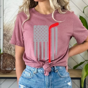 Puede incluir: Una camiseta color malva con un dise&ntilde;o de bandera estadounidense gris y roja desgastada y un palo de hockey rojo. La camiseta est&aacute; anudada a la cintura.