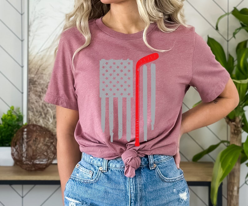 Puede incluir: Una camiseta color malva con un dise&ntilde;o de bandera estadounidense gris y roja desgastada y un palo de hockey rojo. La camiseta est&aacute; anudada a la cintura.