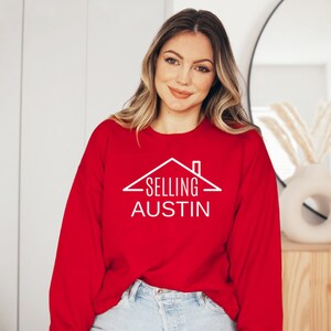 May include: A red sweatshirt with white text that reads "SELLING AUSTIN" and a house graphic above the text.