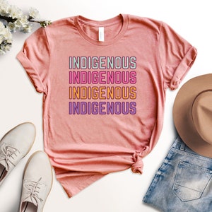 May include: A pink t-shirt with the word "INDIGENOUS" repeated four times in different colors. The word is stacked vertically with each word in a different color: green, pink, orange, and purple.