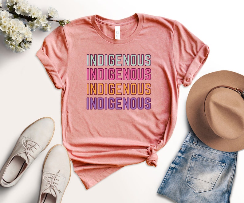 May include: A pink t-shirt with the word "INDIGENOUS" repeated four times in different colors. The word is stacked vertically with each word in a different color: green, pink, orange, and purple.
