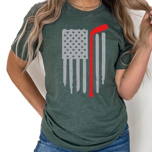 Puede incluir: Una camiseta verde con un dise&ntilde;o de bandera estadounidense desgastada y un palo de hockey rojo. La bandera es blanca con estrellas grises.