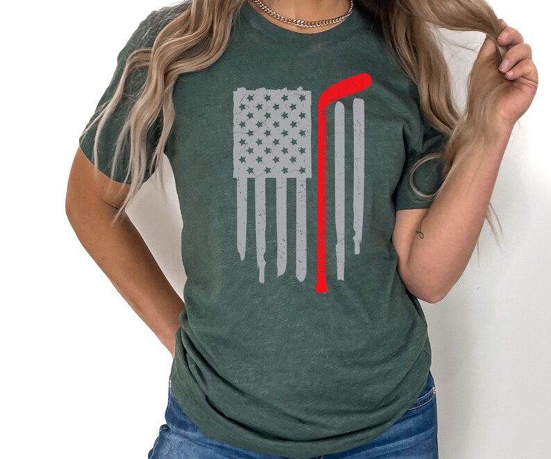 Puede incluir: Una camiseta verde con un dise&ntilde;o de bandera estadounidense desgastada y un palo de hockey rojo. La bandera es blanca con estrellas grises.