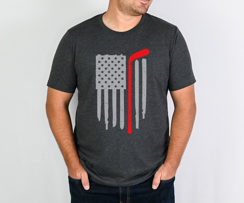 Puede incluir: Una camiseta gris oscuro con un dise&ntilde;o de bandera estadounidense desgastado y un palo de hockey rojo.