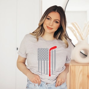 Puede incluir: Una camiseta gris con un dise&ntilde;o de bandera estadounidense roja, blanca y azul. Un palo de hockey se superpone a la bandera. El palo es rojo con una franja blanca.