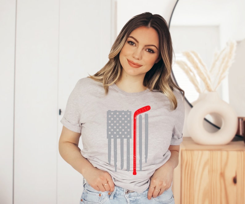 Puede incluir: Una camiseta gris con un dise&ntilde;o de bandera estadounidense roja, blanca y azul. Un palo de hockey se superpone a la bandera. El palo es rojo con una franja blanca.