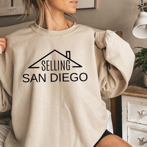 May include: A beige sweatshirt with a black graphic of a house with the text "SELLING SAN DIEGO" on it.