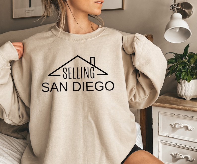 May include: A beige sweatshirt with a black graphic of a house with the text "SELLING SAN DIEGO" on it.