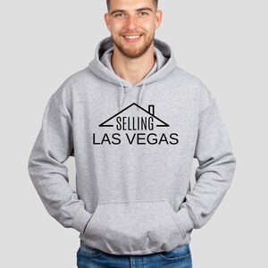 May include: A gray hooded sweatshirt with a black graphic design of a house with the text "SELLING LAS VEGAS" underneath.