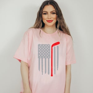 Puede incluir: Una camiseta rosa con un dise&ntilde;o de bandera estadounidense desgastada y un palo de hockey rojo.