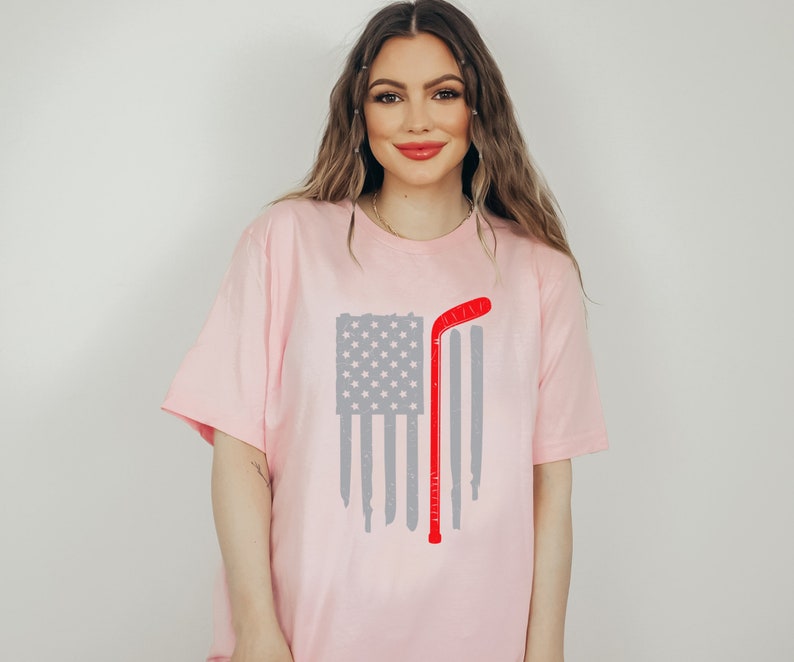 Puede incluir: Una camiseta rosa con un dise&ntilde;o de bandera estadounidense desgastada y un palo de hockey rojo.