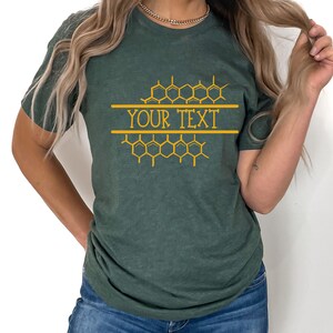 K&ouml;nnte beinhalten: Ein dunkelgr&uuml;nes T-Shirt mit einem Wabenmuster und dem Text "YOUR TEXT" in Gold.