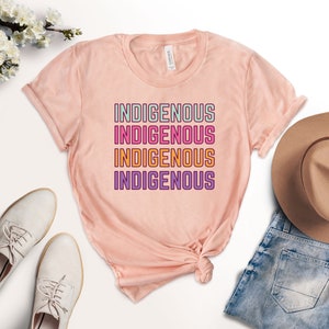 May include: A pink t-shirt with the word "INDIGENOUS" repeated four times in a stacked design. Each word is in a different color: green, pink, orange, and purple.